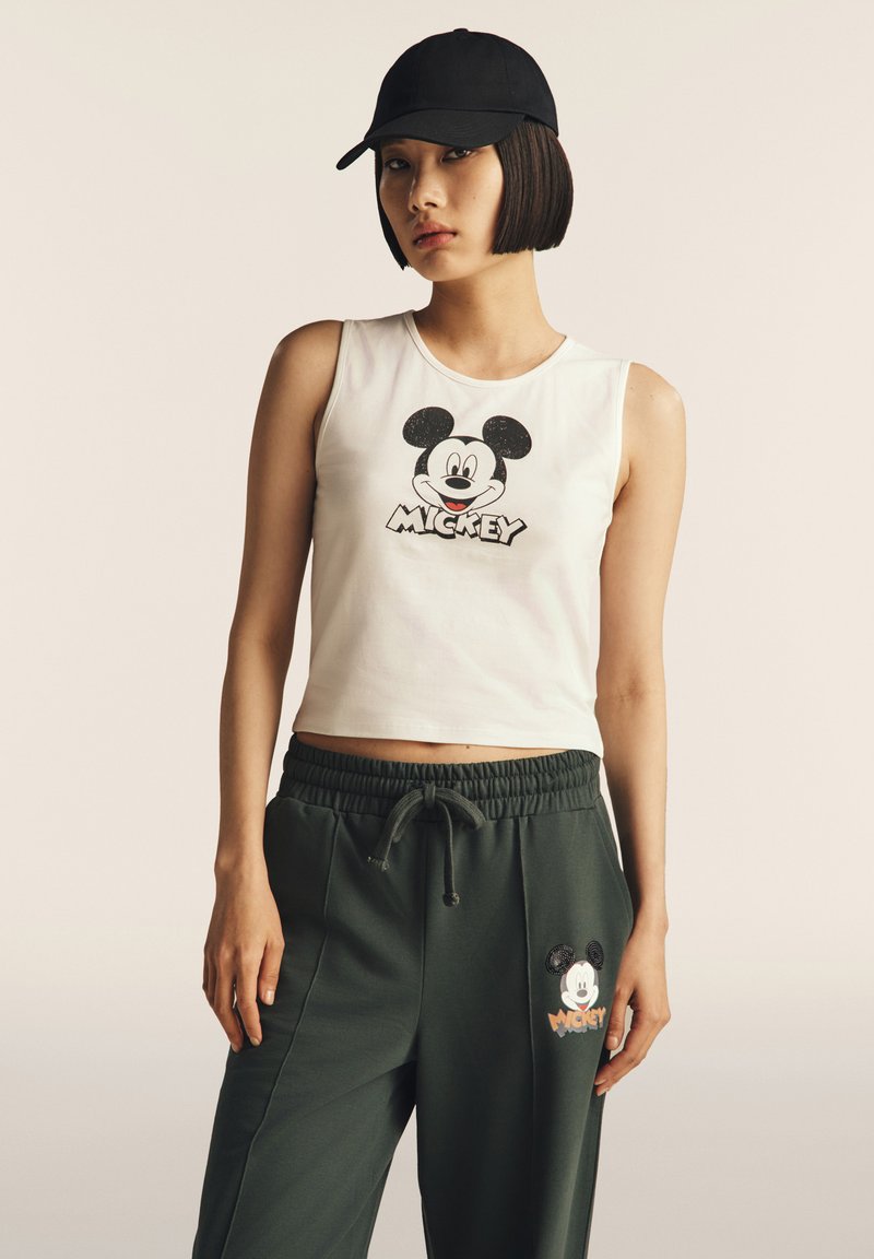 Giovane donna che indossa un cappellino nero, una maglietta senza maniche bianca e pantaloni verde scuro, entrambi con grafiche di Topolino, in piedi su uno sfondo neutro.