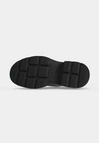Semelle de chaussure noire avec des motifs de bande de roulement texturés rectangulaires et un logo de marque embossé près de la zone de la voûte plantaire, présentée sur un fond clair uni.