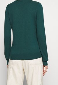 Person som bär en mörkgrön långärmad tröja och off-white byxor, visad bakifrån mot en enkel ljus bakgrund.