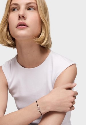 Młoda kobieta z krótkimi blond włosami, ubrana w białą, bezrękawnikową bluzkę, prezentująca na nadgarstku czarną sznurkową bransoletkę z owalnym srebrnym elementem.