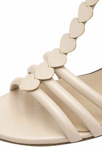 Sandale beige en cuir verni brillant, avec trois larges brides et une chaîne décorative d'accents en forme de cœur.