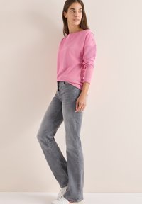 Rosa Strickpullover mit langen Ärmeln und Schulterknopf-Akzenten, kombiniert mit grauen Schlagjeans und weißen Sneakers vor einem neutralen Hintergrund.