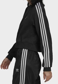 Svart kort sweatshirt i mjukt material med tre vita ränder på varje ärm, tillsammans med svarta byxor med liknande ränder och logotyp.