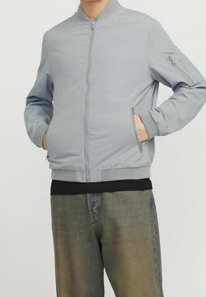 Homme les mains dans les poches, portant une veste bomber gris clair avec poches zippées et un jean délavé vert olive.