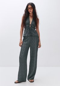 Hauts à col halter vert foncé avec un nœud à l'avant, associés à un pantalon large assorti avec un motif texturé, complété par des lunettes de soleil marron.