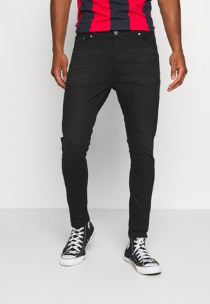 Une personne se tient debout dans un jean skinny noir et des baskets noires, portant un t-shirt rayé rouge et marine, avec un fond blanc uni.