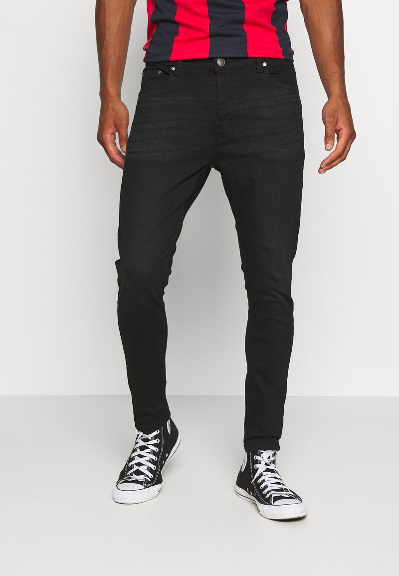 Una persona está de pie con jeans ajustados negros y zapatillas negras, vistiendo una camiseta de rayas rojas y azules, con un fondo blanco liso.