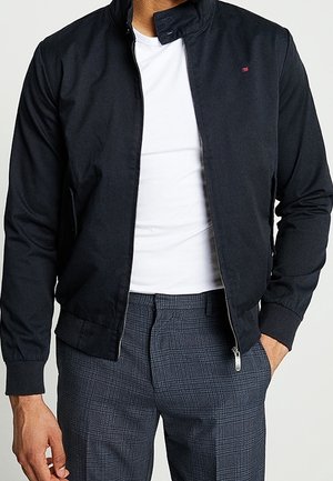 Mann trägt eine schwarze Bomberjacke mit Reißverschluss über einem weißen T-Shirt und grauen karierten Hosen, eine Hand in der Tasche.