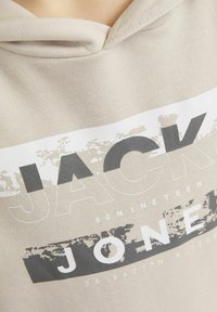 Hoodie beige con stampa grafica che presenta "JACK" e "JONES" in caratteri in grassetto, dettagli bianchi e superficie texturizzata.