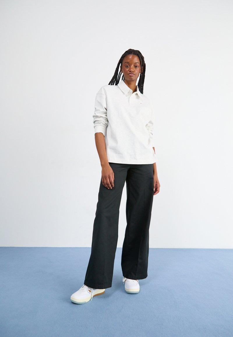 Chemise polo à manches longues gris clair avec col, associée à un pantalon large noir et des baskets blanches avec des semelles colorées sur fond bleu.