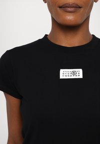 Zwart katoenen T-shirt met korte mouwen, met een wit rechthoekig label met bedrukte cijfers en een cirkelvormig logo op de borst.