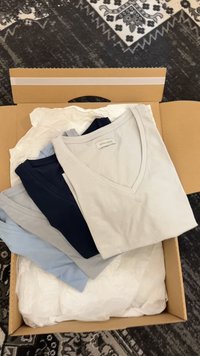 Στοίβα από πέντε διπλωμένα μπλουζάκια με V-λαιμόκοψη σε μπεζ, ναυτικό μπλε, γκρι και ανοιχτό μπλε μέσα σε ανοιχτό χάρτινο κουτί με λευκό χαρτί περιτυλίγματος.