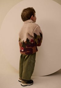 Fuzzy, light pink jacket with multicolored floral and forest graphics, paired with olive cargo pants and brown boots, set against a round backdrop.