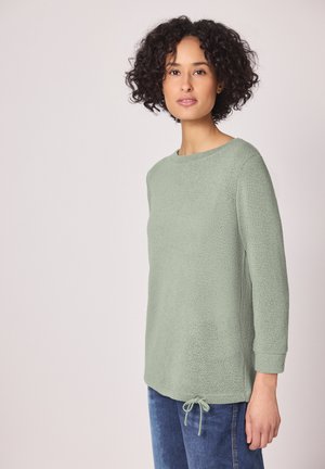 Vrouw met krullend haar, gekleed in een lichtgroene gebreide trui met driekwart mouwen en een trekkoordzoom, gecombineerd met een blauwe spijkerbroek.