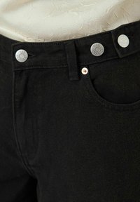 Svarta denimjeans med silverknappar och kopparnitar. Tyget har en lätt struktur och en framficka är synlig.