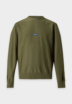 Homme portant un pull vert olive à manches longues, un jean gris foncé, une ceinture noire et des chaussures habillées noires, debout avec une main dans la poche.