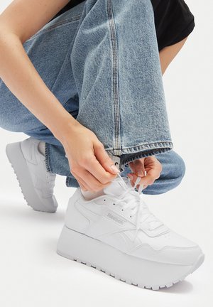 Osoba w jasnoniebieskich dżinsach i białych skarpetach wiążąca białe platformowe trampki Reebok, siedząc na białej podłodze.