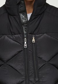 Černá prošívaná bunda s diamantovým prošíváním, dvojitými zipy a kapucí, se značkovým jezdcem zipu a kontrastní texturou.