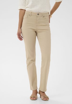 Femme portant un pantalon beige slim, un haut en maille blanc aux manches retroussées, et des sandales à talons ouvertes beige, sur un fond uni.