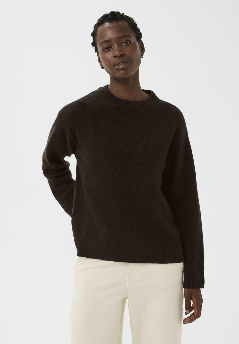 Pulover negru din tricot cu decolteu rotund și manșete și tiv cu un model transparent. Materialul pare moale și confortabil, purtat cu pantaloni de culoare deschisă.
