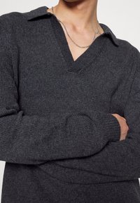 Pull en tricot gris foncé avec un col en V et un col, présentant des poignets côtelés et une finition texturée. Chaîne en argent visible.