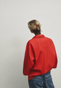 Giacca rossa oversize con colletto a contrasto, tessuto leggero e spalle ribassate, abbinata a jeans in denim blu.