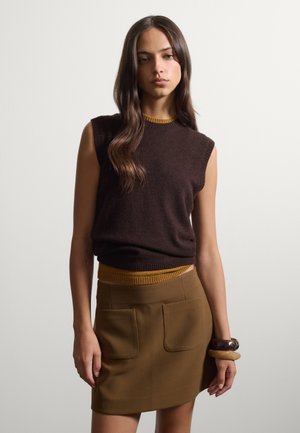Donna con lunghi capelli castani che indossa un maglione senza maniche marrone scuro e una gonna marrone con tasche frontali, in piedi davanti a uno sfondo chiaro e semplice.