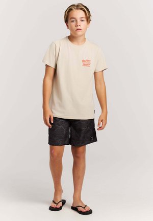 Garçon blond portant un t-shirt beige, un short noir à motifs et des tongs noires, debout devant un fond clair uni.