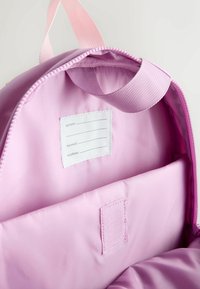 Light pink backpack interior with a fabric loop handle and a white label for name, email, and phone inside the main compartment.