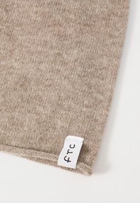 Detailný záber na béžovú pletenú tkaninu s prehnutým lemom a bielou �štítkou z tkaniny s nápisom "FTC" v čiernych písmenách.