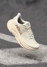 Kremfarget HOKA løpesko med tykk såle, i luften over asfalt med hvit malt linje og skittpartikler under.