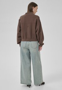 Brown cropped sweatshirt with a high collar, featuring a stitched logo. Paired with light blue wide-leg jeans and black heeled shoes.