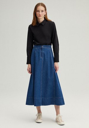 Denim A-line skirt in dark blue with a high waist, button closure, and subtle stitching details, paired with a black collared shirt and beige sneakers.