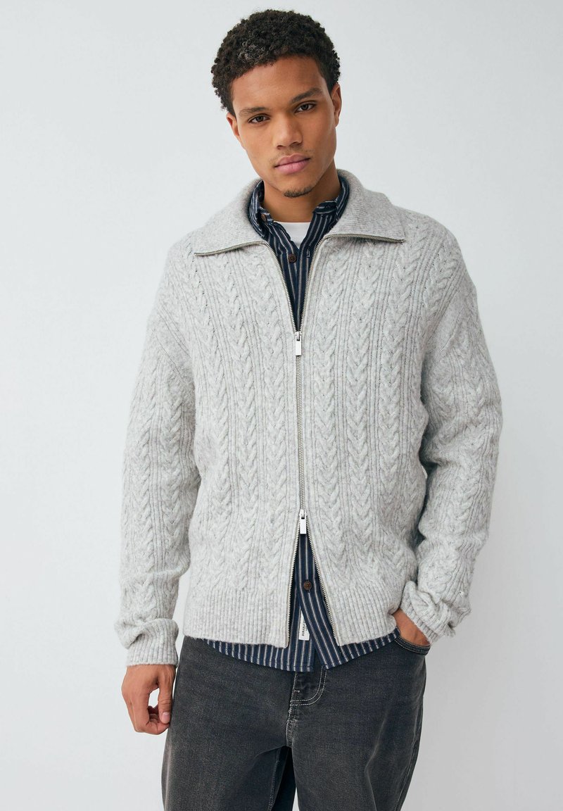 Helehall halli kampsun, mis on varustatud tõmblukuga, kaabelmustriga ja suure kraega. Seljas tumesinise triibulise särgi ja tumedate teksade peal.