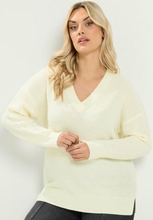 Vrouw met lang blond haar die een oversized crèmekleurige V-hals gebreide trui en donkere broek draagt, staand tegen een effen achtergrond.