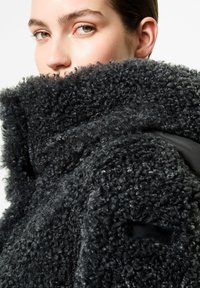 Cappotto in pile nero con texture, caratterizzato da un alto colletto, una morbida fodera interna nera e una piccola tasca decorativa sulla manica.