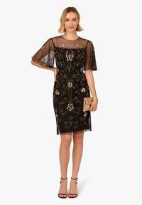 Žena v černých šatech ke kolenům s průhlednými vyšívanými zlatými květinovými vzory, držící zlatou kabelku a oblečená v černých sandálech na kotníky.