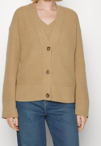 Beżowy dziergany kardigan z głębokim dekoltem w kształcie litery V, ozdobiony trzema dużymi guzikami, ściągaczowymi mankietami i luźnym krojem. Noszony na dopasowanym topie z dekoltem w V.