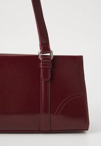 Borsa a mano in finta pelle bordeaux con una forma strutturata, una lunga tracolla regolabile e dettagli in metallo argentato. Presenta una leggera texture e dettagli di cucitura.