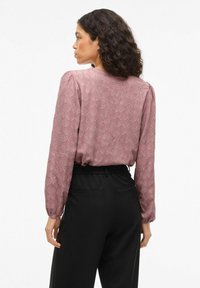 Sieviete ar čirkainiem matiem, tērpusies mauve krāsas teksturētā garajā piedurknē, iebāzta augsto jostasvietu melnās biksēs, skatās pa kreisi uz baltas fona.