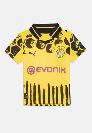 Maillot de football jaune avec un design à motifs noirs et marron, col en V, et logos en violet et noir au centre et sur les manches.