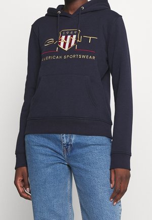 Personne portant un sweat à capuche bleu marine avec le logo doré "GANT 1949 American Sportswear" et un jean bleu clair, debout devant un fond blanc.