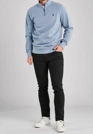 Man draagt lichtblauw sweatshirt met kwart-ritssluiting, zwarte smalle broek en witte sneakers, staat met één hand in de zak tegen een effen achtergrond.