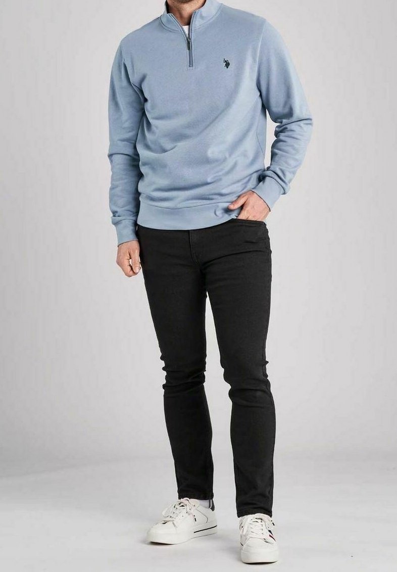 Homme portant un sweat-shirt bleu clair à zip court, pantalon noir slim et baskets blanches, debout avec une main dans la poche sur un fond uni.