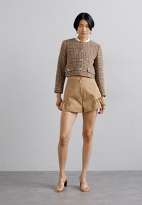 Chaqueta corta de pata de gallo marrón con botones dorados, combinada con pantalones cortos de dobladillo volante en tono beige. La modelo lleva sandalias de tacón transparentes.