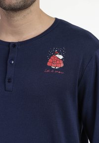 Camisa de manga larga azul marino con una tapeta de botones, que presenta un personaje de dibujos animados con un abrigo rojo y el texto bordado "Que nieve".