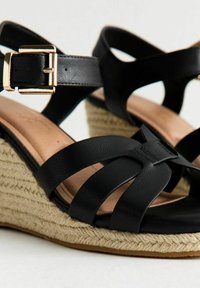 Black leather wedge sandals with crisscross straps, espadrille-style jute detailing, and a gold buckle accent on the ankle strap.