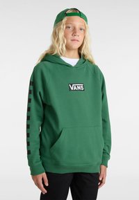 Sudadera verde con un bolsillo delantero y el logotipo de Vans en negro. Presenta un diseño en bloque negro en la manga y una capucha con cordón. Se lleva con una gorra verde a juego.