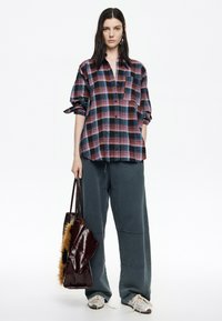 Camisa de cuadros de gran tamaño en rojo, azul y negro, combinada con pantalones de chándal grises holgados. Sosteniendo un bolso burdeos brillante con acento de piel. Zapatillas blancas.