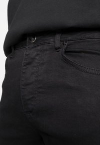 Jeans de mezclilla negra con una textura suave, que cuentan con un bolsillo frontal y un cierre de botón metálico. Costuras visibles a lo largo de las costuras.